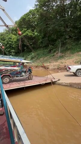This video captures a scene at a river crossing. A ferry, partially visible on the left, is loaded with several motorbikes and a tuk-tuk. A person is operating the tuk-tuk, which is positioned on the ferry. On the left side of the ferry, multiple individuals are seated on motorbikes, some wearing helmets. Another person stands on the ferry near the motorbikes. The ferry is connected to a ramp or dock on the right, with ropes securing it. The river water is a muddy brown color. The background shows a lush green hillside covered in trees, indicating a natural, possibly rural, environment. Overhead, there is a structure with pulleys and cables, likely part of the ferry's operation system. The sky appears cloudy. The camera angle is from a slightly elevated position, looking towards the ramp and the opposite bank. The overall action shows vehicles and people preparing for or in the process of crossing the river via ferry.