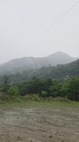 The video shows a misty, overcast day in a rural, mountainous area. The foreground is a dirt, muddy clearing with tire tracks. Behind the clearing, a dense forest covers the rolling hills. The hills are shrouded in mist, and a few small buildings are visible nestled among the trees. A single wire runs diagonally across the frame from the upper left to the upper right. The overall impression is one of a damp, quiet, and slightly melancholic landscape.