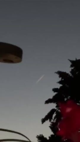 A streak of light moves across the sky, appearing as a faint, elongated glow. The sky is a gradient of dark blue to gray, suggesting it is twilight or dawn. In the foreground, the silhouette of a tree with dark foliage is visible on the right side of the frame. To the left, a portion of a circular object, possibly a light fixture or a piece of equipment, is partially visible. A faint red glow is present at the bottom right corner of the image. The streak of light appears to be moving from left to right and slightly upwards.
