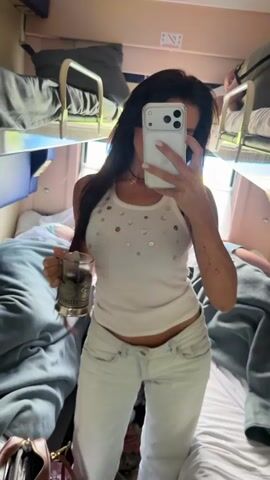 A young woman with long dark hair is taking a selfie in a train cabin. She is wearing a white tank top and white jeans. She is holding a glass with a dark liquid in her left hand. The cabin has bunk beds and luggage. The overall impression is that of a casual, personal moment captured during a train journey.