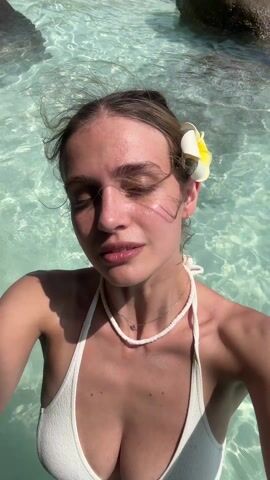 A woman in a white bikini stands in clear turquoise water, taking a selfie. She has light brown hair pulled back with a white and yellow flower tucked behind her ear. She is wearing two necklaces: a white choker and a thin silver chain. The water is calm and reflective, with visible rocks in the background. The sun is shining, casting light on her face.