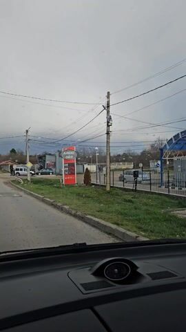 The video shows a driver's view of a road and gas station on a cloudy day. The car is stopped in front of an "АЛЕКС ОИЛ" gas station, and the price signs show the prices of different types of fuel. The road is wet and there are other vehicles parked nearby. There are power lines and utility poles visible. The video begins with the camera pointed towards a gas station sign. The video then shows the road ahead through the windshield of a car. There appears to be a rainbow in the distance. A few people are visible on the side of the road.