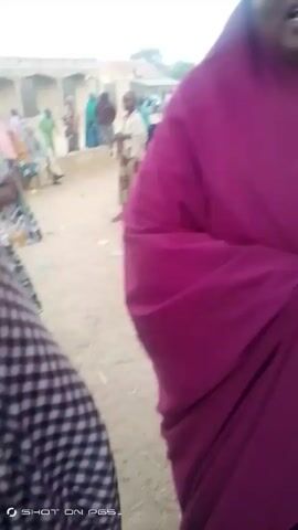 The video shows two women talking, wearing head coverings, in a town or village. They are standing in an open area with other women and children sitting or standing around them. The background includes buildings and trees. One woman is wearing a solid red head covering and the other is wearing a red and black checkered covering. The video is shaky and the camera angle is low. The video appears to be shot on a phone.