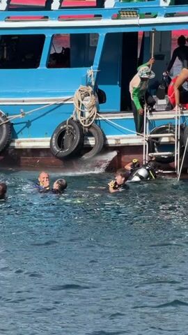 A blue boat with a red stripe along its side is docked in clear blue water. Two large black tires are attached to the side of the boat. A rope is coiled on the deck of the boat, near the tires. There are several people in the water surrounding the boat. Some of them are wearing scuba diving gear. One person is climbing a ladder onto the boat. Another person is standing on the deck of the boat, wearing a hat and a green shirt. The person on the deck is holding a blue bag and a red life preserver. The water is choppy.