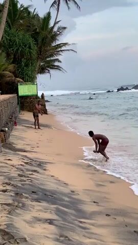 Two shirtless men are playing catch with an object on a beach near the ocean. The sand is light with dark streaks. Palm trees and vegetation are on the left side, next to a wall. A green information sign is present. The ocean has waves breaking near rocks, and people are visible in the water. The sky is overcast.
