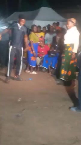 The video shows a group of people gathered outdoors at night, possibly for an event or celebration. There are tents in the background, suggesting a temporary setup. In the foreground, several individuals are dancing. One young man in a black tracksuit with white stripes is actively dancing, with his body angled and his arms in motion. Another person, wearing a colorful patterned shirt, is also dancing energetically in the center of the frame. Many of the women in the crowd are wearing brightly colored skirts with distinct patterns, and some are seated. The ground appears to be dirt. The lighting is dim, indicating it is nighttime, and there are visible light sources that illuminate the scene.