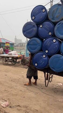 The video shows two men pushing heavily loaded carts on a dirt road near a river. In the first scene, a man is standing next to a cart loaded with blue plastic barrels, his hand resting on one of the barrels. He wears a brown shirt, a patterned green and brown sarong, and orange sandals. He has short dark hair. In the second scene, another man in a striped t-shirt and colorful sarong is pushing a cart loaded with cardboard boxes. He has short dark hair and is leaning into the cart, using his weight to help push it. The carts are on a dirt road alongside a river. There are other carts and bicycles visible in the background, along with buildings on the opposite side of the river. The sky is overcast.