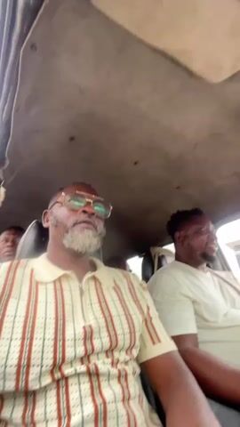 The video shows the interior of a vehicle from a first-person perspective. Two men are visible. The man in the foreground on the left is wearing glasses and a striped shirt. He is looking to the left. The man in the background on the right is wearing a white t-shirt and is sitting in the driver's seat, looking forward. The ceiling of the vehicle is visible above them. The lighting suggests it is daytime.