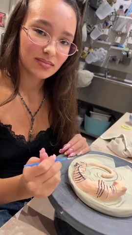 A young woman with long, dark hair and pink-rimmed glasses is sitting at a pottery wheel, painting a clay sculpture of a curled-up tiger with black stripes. She is wearing a black lace-trimmed tank top and a blue beaded necklace. Her left hand holds a small paintbrush, and her right hand is near the sculpture. The sculpture is on a gray pottery wheel, and it appears to be a light brown color with black stripes applied to it. The background shows a workshop environment with shelves filled with pottery supplies and tools, including sinks and various containers.