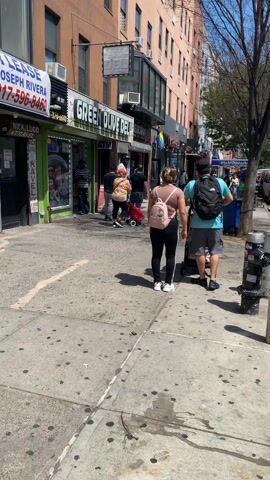 The video captures street scenes in New York City on a bright, sunny day.

The initial segment shows a busy sidewalk lined with storefronts. In the foreground, a woman with a pink backpack and black pants stands beside a man in a blue shirt, gray shorts, and a black backpack, who is pushing a baby stroller. Several other pedestrians are seen walking. Visible businesses include "GREEN OLIVE DELI" and a small "CHIPOTLE" sign. A partial phone number "370-6640" is visible on a sign.

The scene then shifts to the street, revealing parked cars and a white NYPD police SUV. The police vehicle, marked "4441" and "25 PCT" with "COURTESY PROFESSIONALISM RESPECT" and "NYPD" on its side, has its red and blue lights flashing. Its driver's side door is open. A blue car with a New York license plate "MBY-9196" is parked in front of the police SUV. In the background, "T-MOBILE AUTHORIZED RETAILER" and "POPULAR" bank signs are clearly visible, along with "ELITE NAILS SALON" and the phone number "917-388-3143."

The video concludes with another view of the sidewalk, showing more pedestrians, parked cars, a tree, and a blue trash can. The overall setting is a vibrant urban environment with commercial establishments and ongoing daily activity.
