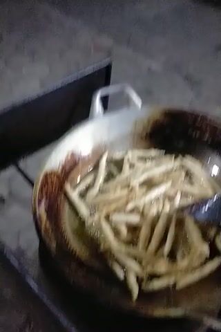 The video shows a close-up of a pan filled with french fries, with oil. In the background, there are motorcycles and people standing around. The lighting suggests it is nighttime or indoors. The scene appears to be an outdoor food stall or street market.