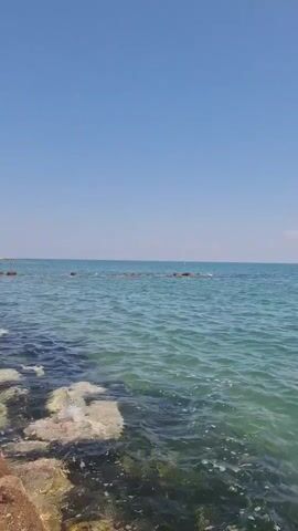 The video shows a serene view of the sea under a clear blue sky. The water is a beautiful shade of turquoise, with gentle waves lapping at the shore. The foreground is occupied by rocks and some submerged structures, while the horizon stretches out to meet the sky, dotted with a single, small cloud. The overall atmosphere is peaceful and calming.