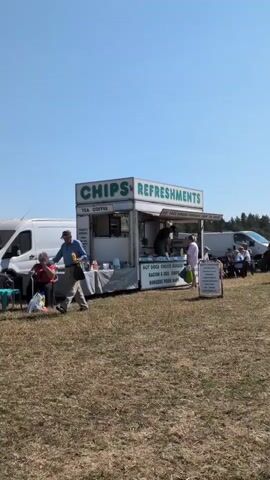 The video shows a food stand labeled "CHIPS REFRESHMENTS" in an outdoor grassy area under a clear blue sky. A person in a green apron is seen working behind the counter, preparing food. A customer, a woman wearing a pink top and light-colored pants, is standing at the counter, possibly ordering. A sign board next to the stand displays a menu with items like "HOT DOGS", "CHEESE BURGERS", "BACON & EGG", "CHIPS", "BURGERS", and "PORK BAPS". In the background, several people are seated at a table to the right, and vans are parked to the left and right of the food stand. A man in a light blue shirt and khaki pants is walking away from the food stand towards the left side of the frame. The ground is covered in dry, brown grass.