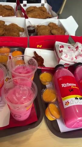 The video shows a close-up, slightly overhead view of a table laden with food and drinks. Dominating the frame are several red and white cardboard boxes containing fried chicken pieces. In front of the chicken boxes, two silver trays are visible. The tray on the left holds four clear plastic cups filled with a pink, icy beverage, and several small containers of orange dipping sauce. The tray on the right contains more orange dipping sauce containers, a white paper container with what appears to be a few glazed chicken wings, and a large pink bottle labeled "Richeese Factory." Several other smaller items, like crumpled paper napkins and receipts, are also present on the table. The overall impression is of a large, shared meal, likely from a fast-food restaurant. The lighting suggests an indoor setting, possibly a restaurant or home. No people are directly visible, but the presence of multiple servings implies a group dining.