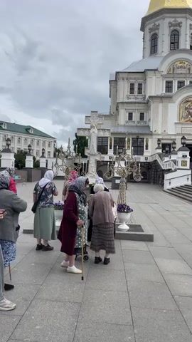 A group of people, mostly older women wearing headscarves, are gathered around a large white cross in front of a large, ornate white building with a golden dome. The building appears to be a church or a similar religious structure. Some of the women are holding canes and appear to be praying or touching the cross. One younger person, a teenage boy, walks past the group. The ground is paved with gray stones, and there are white planters with purple flowers. The sky is overcast.