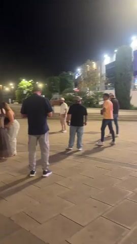 This is a nighttime video taken in Alcorcón, Spain. The scene appears to be an outdoor pedestrian area, possibly a plaza or a walkway in front of shops. The lighting suggests it is evening or night.

Several people are visible in the foreground and background. In the foreground, three men and two women are standing or walking. One man in the very front is wearing a dark blue shirt and grey cargo pants. Further back, a man in a white hat and beige pants stands in the middle, while another man in a black t-shirt and blue jeans is to his right. Two women are also present, one in a patterned dress and the other in light-colored pants and a dark top.

In the background, a woman in white pants and a black top is seen walking and then running across the plaza. A building with multiple businesses is visible behind her. Signs on the building indicate several establishments: "100 Montaditos," "Pomodoro," "Rodilla," and "Pans & Company." The architecture of the building suggests a modern shopping or dining complex.

The overall atmosphere seems relaxed and casual, with people gathered or passing through the area. The ground is paved with large square tiles, and there are some trees and outdoor seating areas visible. The time of day is clearly night due to the artificial lighting from the buildings and the dark sky.