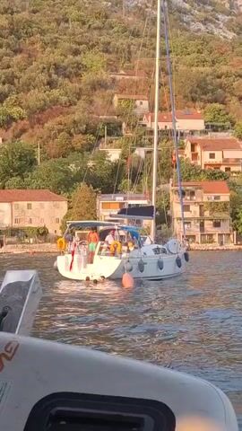 A white sailboat with a mast and sails is docked in the water. People are on the deck of the sailboat and in the water. Some people are swimming and playing in the water near the sailboat. There are buildings and trees on the hillside behind the sailboat. The water is calm and the sun is shining. The image is taken from the perspective of another boat in the foreground, which is partially visible.