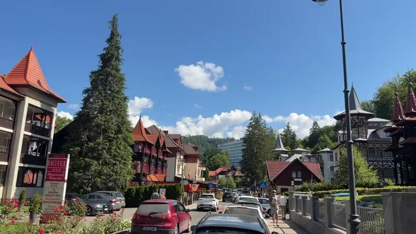 The video shows a street in a town with buildings and trees. There are cars parked and driving on the street. Some people are walking on the sidewalk. The sky is blue with some clouds. There are hotels and traditional-looking buildings with red roofs. In the background, there is a large white hotel with the word SOVATA on it. The scene appears to be a daytime shot on a sunny day.