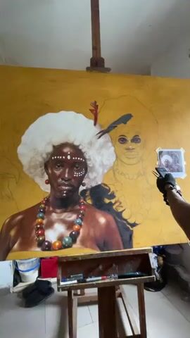 A close-up shot displays an art canvas with two figures in progress. The figure on the left is further along, detailed with brown skin, white face paint, and a beaded necklace, topped with a large white afro wig and a feather. To the right, a figure is still in the sketched phase with minimal detail, also sporting an afro. A reference photograph is taped to the upper right corner of the canvas. An easel supports the canvas, and an artist is captured applying paint to the piece using an airbrush-style tool. The environment suggests an art studio or workspace.
