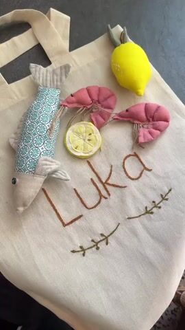 A close-up, high-angle shot shows a canvas tote bag with the name "Luka" embroidered on it in brown thread. Various plush toy food items are arranged on the bag. To the left, a plush fish toy is positioned vertically. In the center, there is a plush lemon slice, two plush shrimp toys, and a plush lemon toy. Additional plush food items, including a carrot, avocado, tomato slices, cheese slices, mushrooms, broccoli, a cucumber, a banana, a burger bun, and a sausage, are piled on top of and around the central area of the bag, obscuring part of the name. Some of the plush food items have visible stitching and stuffing. The background is a dark, textured surface. The lighting is bright, highlighting the colors and textures of the plush toys.