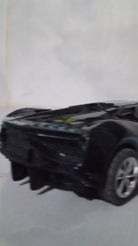 This is a short video showing a black toy car from the side. The car is parked on a flat surface, possibly a table or floor, with a light-colored wall in the background. The lighting is dim, creating shadows. The camera moves slightly, giving a brief glimpse of the car's design, including its wheels and body.