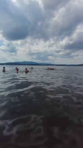The video begins with a low-angle shot of a body of water on a cloudy day. In the distance, several people are swimming and one person is lying on a paddleboard. The water is choppy, with visible waves. Hills or mountains are visible in the far background, along with a hazy horizon. The sky is filled with dramatic, dark clouds, suggesting an impending storm or recent rain.