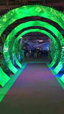 The video shows a wide-angle view of an event space, likely a hall or convention center. In the foreground, there is a red carpet leading into the space, flanked by arches covered in green LED lights displaying a leafy pattern. Tall green plants in black pots are placed on either side of the arches. As the camera moves forward, the scene opens up to reveal the main event area. There are round tables with stools and high-top tables with stools scattered throughout the space. A stage is visible in the distance with a band or performers on it, illuminated by stage lights. Several people are standing and sitting at the tables, and some are walking around. The overall atmosphere appears to be a festive gathering or party.