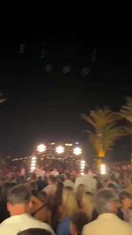 A crowd of people are gathered at night, illuminated by warm-toned lights. In the foreground, the backs of several individuals are visible, facing a stage area. The stage is adorned with multiple light fixtures, some resembling clusters of bulbs and others more industrial in design. A palm tree is visible in the background, silhouetted against the dark sky. People in the crowd are looking towards the stage, and some appear to be holding up their phones, likely to record or photograph the event. In the middle ground, there is a person playing a woodwind instrument, possibly a clarinet, and another individual standing behind a console, possibly a DJ. The overall atmosphere suggests an outdoor concert or performance event.