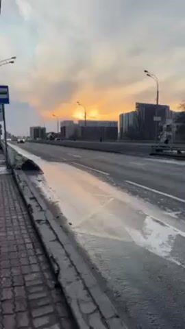 The video shows a roadside scene with a multi-lane road in the foreground and buildings in the background under a sky with a bright sunset. The road is wet, and some patches of white paint or residue are visible. A silver car passes by on the road, moving from left to right in the frame. There's a bus stop sign on the left side of the screen. The sky is filled with clouds illuminated by the setting sun.