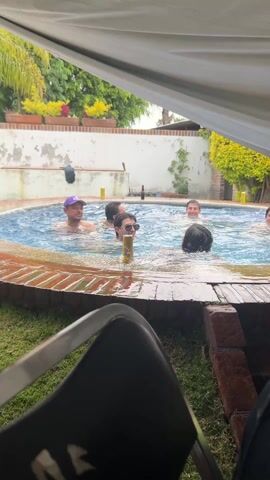 The video shows a group of about seven people in a small pool in a backyard setting. The pool has a distinctive brick border and blue water, with several beer cans floating around or near the people. The people are all mostly submerged, suggesting they are relaxing or swimming. One person wears a purple baseball cap, while another has sunglasses. They seem to be socializing. The backyard includes a wall with potted plants and greenery. The video is shot from under some sort of canopy or awning, with a black plastic chair partially visible in the foreground. The overall atmosphere suggests a casual, social gathering, likely during the daytime.