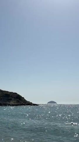 A vertical video opens to a bright, clear blue sky over a vast expanse of shimmering blue ocean. In the distance, a small, dark, dome-shaped island is visible on the horizon. To the left, a rocky, sparsely vegetated hill slopes down to the water's edge. The sea is calm with gentle waves, and the sun's rays create sparkling reflections on the surface. There is a brief, barely visible appearance of a person swimming in the lower right corner of the frame around the 00:01 mark.