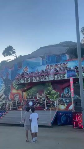 The video opens with a wide shot of an Avengers-themed amusement park ride. The ride is adorned with large images of Hulk, Iron Man, Captain America, and other Avengers characters. A group of people are seated on the ride, which appears to be in motion, their arms raised in the air. Two young men, one in a beige t-shirt and shorts, the other in a white t-shirt and shorts, stand facing the ride, looking up at it. The background shows a grassy hill with some trees and a clear blue sky. The ground in front of the ride is sandy or dirt. There are lights on the ride and on a small structure to the right. The ride itself has a raised platform with stairs leading up to it.