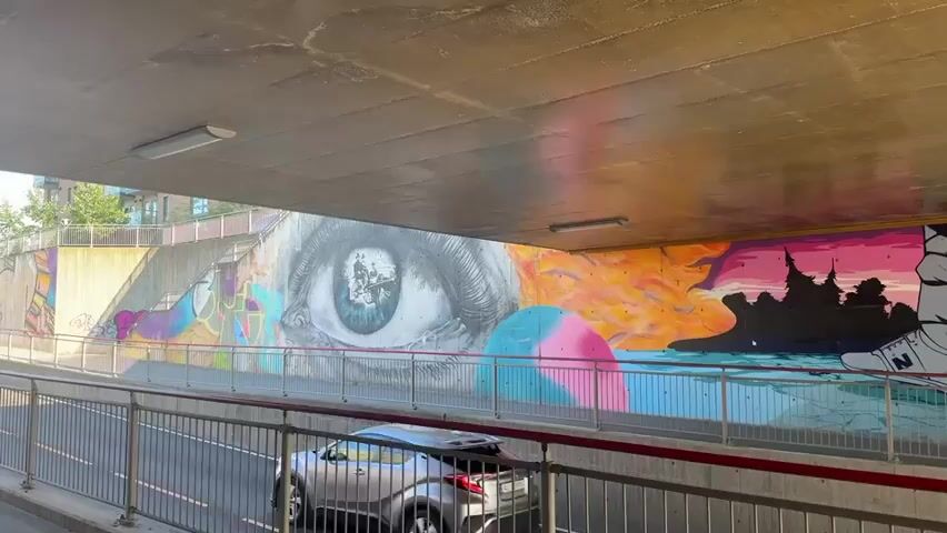 A car drives past a mural on the wall of an underpass. The mural depicts a giant eye looking out from the left side. To the right of the eye, abstract shapes in orange, yellow, pink, and blue are visible. Further to the right, a row of legs and feet wearing shoes and boots are depicted. The legs are walking on a blue surface that appears to be water or a reflection. In the background, a silhouetted city and a pink and purple sky are painted. The underpass has a concrete ceiling with fluorescent lights and a concrete wall with metal railings along the walkway.