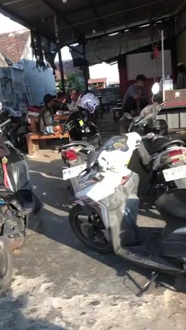 The video shows an outdoor gathering at what appears to be a roadside eatery or rest stop. Several motorcycles are parked in the foreground, suggesting transportation is a key element. In the background, a group of people are seated at tables, some interacting with each other, and one person is on a motorcycle, wearing a helmet. The environment is rustic, with visible roof structures and outdoor seating. The lighting suggests it is daytime.