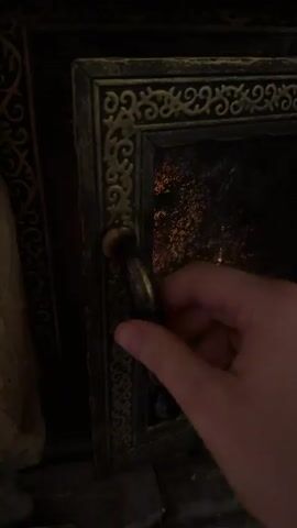A person opens the door to a wood-burning stove, revealing a lively fire. The stove is made of dark metal and has decorative trim. The fire consists of burning wood logs resting on a bed of glowing embers. The interior of the stove is brick. The door is closed again, leaving the fire burning inside.