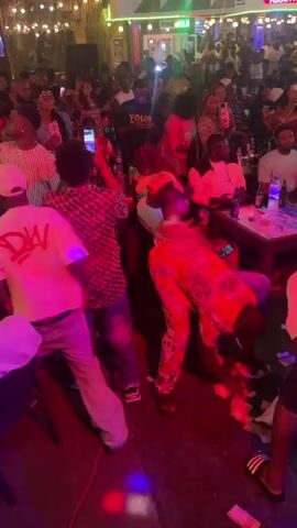 The video shows a crowded outdoor venue at night. The lighting is a mix of ambient light from the surroundings and colorful stage lighting, primarily pink and blue. Many people are present, appearing to be socializing and enjoying themselves. Some individuals are standing, while others are seated at tables. There are drinks and possibly food on the tables. Several people are holding up their phones, suggesting they are recording or taking pictures. At least one person is bending down and picking up items from the ground, which appear to be bills or papers. The overall atmosphere suggests a lively social gathering or event.