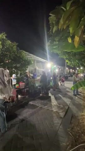 The video captures a nighttime scene at what appears to be an outdoor food stall or small market. Several people are gathered around a stall with a bright light illuminating the area. The stall has tables and what seems to be food being prepared or displayed. The ground is paved with bricks, and there are trees and foliage on either side of the walkway. The overall atmosphere suggests a casual, public gathering place at night, possibly a street food vendor or a small evening market.