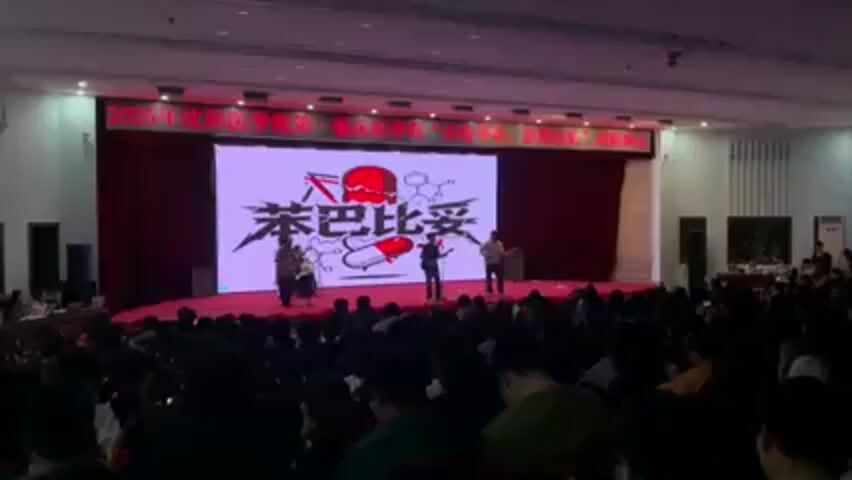An indoor auditorium is shown with a band performing on a stage in front of an audience. The stage has a large screen displaying text, a stylized skull, and depictions of chemical compounds. The band consists of at least three musicians playing instruments, including guitars and drums. The audience is seated and appears to be watching the performance. The room is well-lit with a red curtain hanging behind the screen. The band is performing rock music.