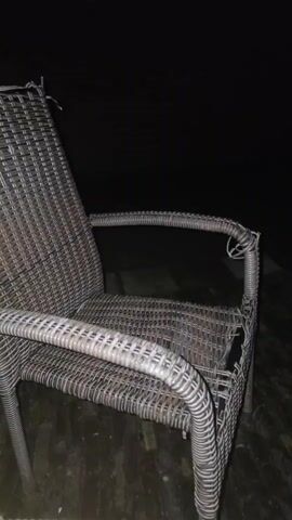 The video begins by showing a wicker chair in what appears to be an outdoor setting at night. The chair is made of woven material and has a metal frame. The camera then pans to the right, revealing a dark, indistinct background. The lighting is very low, making it difficult to discern details of the environment. The focus remains on the chair and the darkness beyond. The video ends with the camera still pointed towards the dark background.