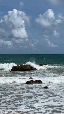 A video shows waves crashing against rocks in the ocean. The sky is blue with white clouds. The water is turquoise and there is white foam from the waves. The rocks are dark brown and covered with some green moss or algae. There are several rocks visible, some larger and some smaller, with waves breaking around them. The foreground shows a sandy beach with some shallow water and foam. The video is shot from a stationary perspective at a low angle.