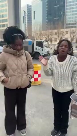 Two Black women stand outside on a sidewalk, facing the camera, waving. The woman on the left wears a tan puffer jacket and the woman on the right wears a black and white sweater. Both women wear dark pants and dark shoes. Behind them is a street with cars, a traffic barrier, and high-rise buildings. The day appears overcast.