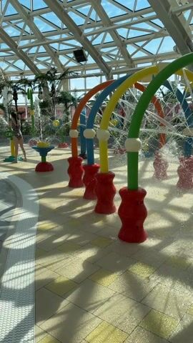 The video shows an indoor water park area with colorful water arches spraying water. Two young girls in swimsuits are playing in the water area. The building has a high, lattice-style glass roof letting in natural light. There are decorative palm trees visible in the background. The water area has a tile floor. The arches are red, blue, yellow, and green. Water sprays from the arches, creating a playful atmosphere.