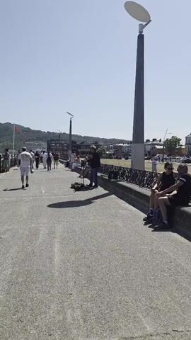 The video captures a bright, sunny day with a clear blue sky. A paved walkway runs parallel to a body of water, though the water itself is not visible. A low, decorative railing lines the edge of the walkway near the water. On the walkway, a lone musician is playing an instrument, possibly a guitar, and singing into a microphone that is on a stand. They are seated on a low wall or bench. Several people are walking along the walkway, and others are sitting on benches along the railing, watching or listening to the musician. In the background, there are buildings, trees, and a hill in the distance. Tall, modern streetlights are also visible along the walkway.