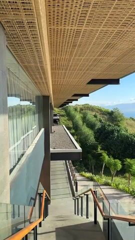 The video shows a high-angle view of a modern building overlooking a large body of water. The building has a flat roof covered in a woven pattern. A set of stairs with wooden handrails and glass panels leads down the side of the building, along a landscaped pathway. The view from the building looks out over a green hillside with trees towards a wide, calm body of water under a clear, blue sky with some clouds. The sun is shining brightly, reflecting on the water. There are distant landmasses visible on the horizon, and a few small boats can be seen on the water. The overall impression is one of a luxurious and scenic location. A very loud droning sound is constant.