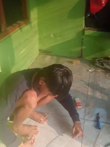The video shows a top-down view of two young boys, likely children, sitting on a tiled floor. They are in the process of constructing a kite. One boy, closer to the camera, is shirtless and is using his feet to hold down some kite string while his hands manipulate other strings. He is wearing a dark long-sleeved shirt with a yellow collar. The other boy, further away, is wearing a long-sleeved dark shirt and is also engaged in the kite-making process, holding onto a part of the kite frame. Various materials related to kite construction are scattered around them, including bamboo sticks, string, a roll of paper or plastic (purple in color), a small knife or cutter, and a yellow object that might be tape or a lighter. The environment appears to be indoors, possibly a room or a covered outdoor area, with green walls visible on the left side. The lighting suggests it is daytime. The boys are actively working on the kite, demonstrating cooperation and engagement in the activity.