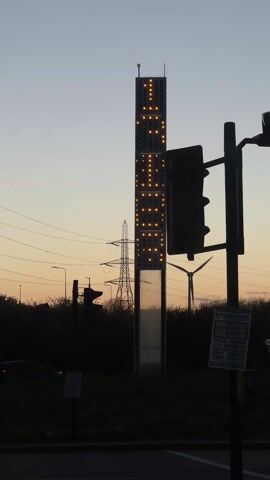 The video shows a vertical tower with lights on it, forming the word "LEEDS".  The tower is located near a road with traffic lights, and in the background, there are power lines and a wind turbine. A poster for the "51st State Festival" is attached to the traffic light pole. The sky is a dusk color.