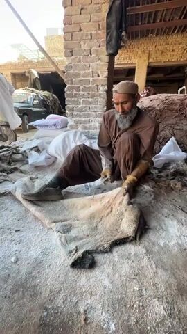 An elderly man with a long gray beard and a tan cap sits cross-legged on a concrete floor covered in white powder. He wears a brown outfit, gloves, and dark-colored shoes. He is working on a large, light-colored animal hide, using a tool to scrape or clean its surface. The hide has dark patches around its edges. Behind him, a brick pillar supports a roof structure. In the background, there are bags of white material, a vehicle, and other people moving around. Piles of what appears to be more animal hides are stacked in the background. The lighting suggests an outdoor or semi-outdoor workspace. The scene appears to be a traditional leatherworking environment.