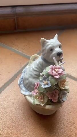 The video shows a white porcelain figurine of a dog sitting in a basket overflowing with flowers. The dog is a West Highland White Terrier with its ears perked up. It has a blue collar around its neck. The basket is decorated with a light blue cloth. The flowers are in various pastel shades of pink, purple, and white, with green leaves. A small white butterfly is perched on one of the flowers. The figurine is placed on a terracotta tiled floor. The background consists of a dark brown wooden surface with horizontal lines. The lighting is natural and appears to be coming from the side, casting a slight shadow on the floor. The video is a close-up shot of the figurine, with the camera slowly zooming out. The timestamp indicates the video duration is approximately 4 seconds. There are no people or any other living beings visible in the video. The scene is static, with no movement except for the subtle camera zoom.