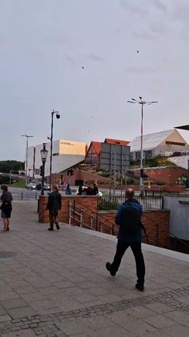 The video shows a street scene with several people walking. In the foreground, a man wearing a blue jacket and backpack walks away from the camera on a paved sidewalk. To his left, a woman with a backpack and blonde hair stands leaning on a low wall. Further left, a woman in a pink shirt and black leggings walks away from the camera. In the background, there are several modern buildings with large windows and geometric designs, some with brick accents. A street light is visible on the left side of the frame, and a traffic light is also present. Several red bicycles are parked in a row on the right side of the sidewalk. The sky is overcast with some clouds. The overall impression is an urban environment during the day.