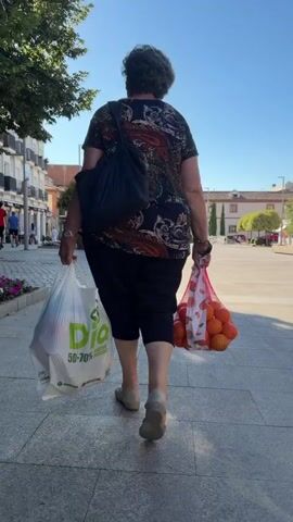 This video shows a woman walking away from the camera on a paved walkway. She is carrying two bags: a white plastic bag with green text on her left and a pink mesh bag filled with oranges on her right. She is wearing a dark patterned shirt, black capri pants, and light-colored shoes. A dark blue bag is slung over her left shoulder.

The scene is set outdoors on a sunny day, indicated by the strong shadows cast on the ground and the clear blue sky. The background features buildings with balconies, trees, and a lamppost, suggesting an urban or suburban environment. The architecture and the visible signage imply the location is Las Rozas de Madrid, Spain.

The woman appears to be returning from grocery shopping, based on the contents of her bags. Her gait suggests a steady, purposeful walk. The overall mood is ordinary and peaceful, depicting a common daily activity. The visible text on the white bag includes "Dia 50-70% Reciclable," which is a supermarket chain in Spain known for its discount prices and emphasis on recycling.